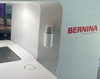 May include: A white sewing machine with a black control panel and a large screen displaying the letters "ABC". The machine has a red light on the front and the word "BERNINA" printed on the side.