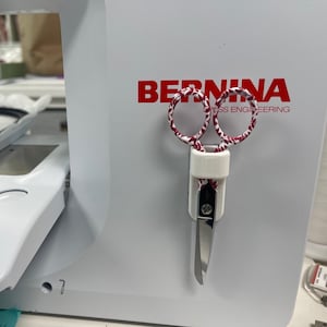 May include: White sewing machine with a red and white patterned pair of scissors attached to the side. The machine has the word "BERNINA" and the text "Swiss Engineering" printed on the side.