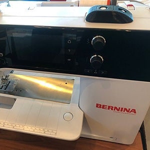 May include: A white sewing machine with a black digital display and a BERNINA logo. The machine has a needle and a foot pedal.