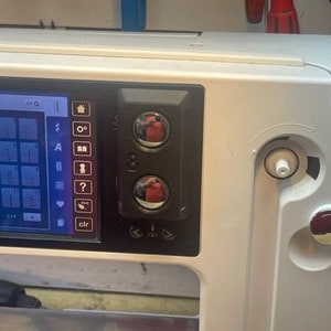 May include: Close-up of a white sewing machine with a digital display showing a variety of stitch options. The machine has two red buttons and a white knob.