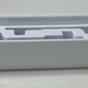 May include: White plastic needle plate holder with the text "45789 Needle Plates Holder" printed on the top.