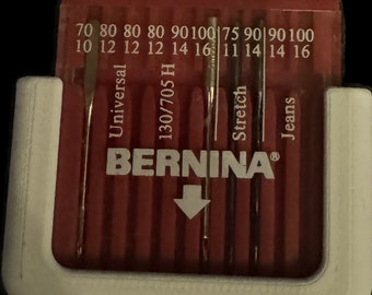 May include: A red plastic case with a variety of sewing machine needles. The needles are labeled with different sizes and types, including Universal, 130/705 H, Stretch, and Jeans. The case also has the BERNINA logo and a white arrow pointing down.