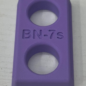 May include: A rectangular, purple object with two circular holes. The text "BN-7S" is embossed on the front. The object has rounded edges and a smooth surface. The item appears to be 3D-printed.