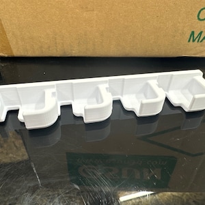 May include: White plastic organizer with five slots for holding items. The organizer is designed to be mounted on a wall.