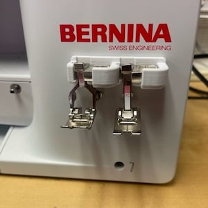 May include: A white sewing machine with the brand name "BERNINA" and the text "SWISS ENGINEERING" printed on the front. Two silver sewing machine feet are attached to the side of the machine.