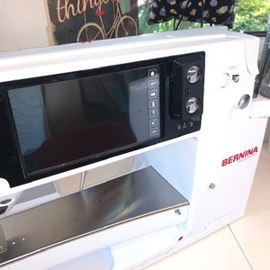May include: White sewing machine with a large black screen and a red BERNINA logo. The screen has a menu of options and a digital display.