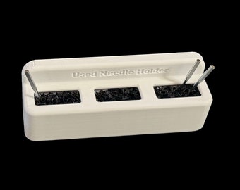 May include: White plastic needle holder with three compartments filled with black needles. The holder has the text "Used Needle Holder" printed on the front.