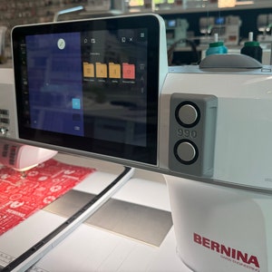 May include: A white Bernina 990 sewing machine with a large touchscreen displaying file folders. The machine is stitching a red fabric with a white pattern. The machine has a gray control panel with two buttons.