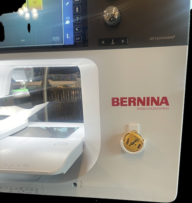Op de afbeelding: Een witte naaimachine met een zwart-wit scherm. De machine heeft een rood "BERNINA" logo met de tekst "SWISS ENGINEERING" eronder. Een klein geel en wit accessoire is aan de zijkant van de machine bevestigd.
