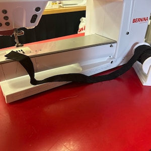 May include: White sewing machine with a black fabric strap attached to a white spool holder. The machine is on a red surface.
