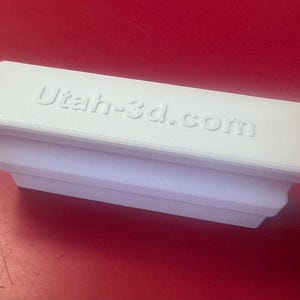 May include: A white rectangular 3D-printed object with the text "Utah-3d.com" embossed on the top. The object has a stepped design and sits on a red surface. The item appears to be a custom-made product.