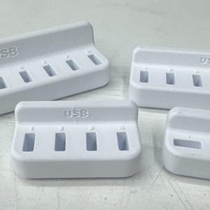 May include: Four white USB charging stations of varying sizes are arranged on a light surface. Each station has multiple rectangular slots for charging devices. The word "USB" is printed on each station.