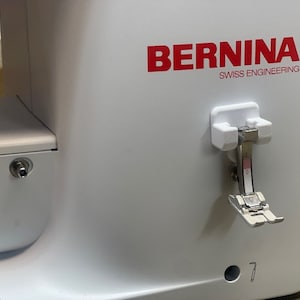 May include: A white sewing machine with the BERNINA logo and the words "SWISS ENGINEERING" printed in red. The machine has a white foot pedal attached to the side.
