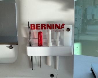 May include: A white sewing machine with a white accessory holder attached to the side. The holder has a red screwdriver, a white needle, and a white tool inside. The sewing machine has the word "BERNINA" printed on the front in red letters.