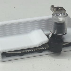 May include: A white 3D-printed sewing machine attachment with a black and silver blade. The attachment has a clear plastic cover and a metal clamp. The device is designed to assist with sewing tasks.