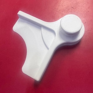 May include: A white, 3D-printed object with a unique shape. It features a rounded top, a handle-like extension, and a curved section. The object is made of a ridged material and sits on a red surface.