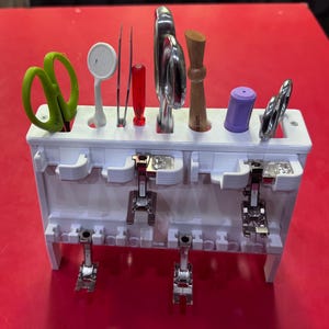 May include: A white 3D-printed sewing tool organizer with various sewing tools, including green scissors, tweezers, and sewing machine presser feet. The organizer is on a red surface.
