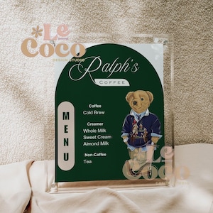 May include: A clear acrylic menu holder with a green background featuring the text "Ralph's Coffee". The menu lists coffee, cold brew, creamer, milk, and tea options. A teddy bear wearing a navy blue sweater is also featured.