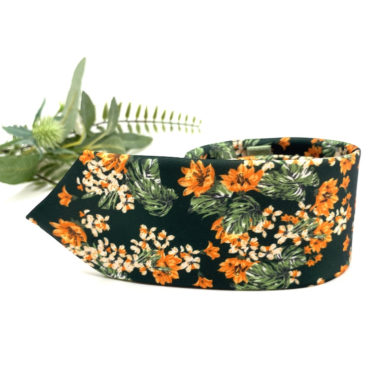 Peut inclure: Une cravate vert fonc&eacute; orn&eacute;e d'un motif r&eacute;p&eacute;titif de fleurs orange vif et de feuilles tropicales vertes. Le tissu pr&eacute;sente une texture lisse et la cravate est pli&eacute;e en forme circulaire, mettant en valeur l'imprim&eacute; floral sur toute la surface.