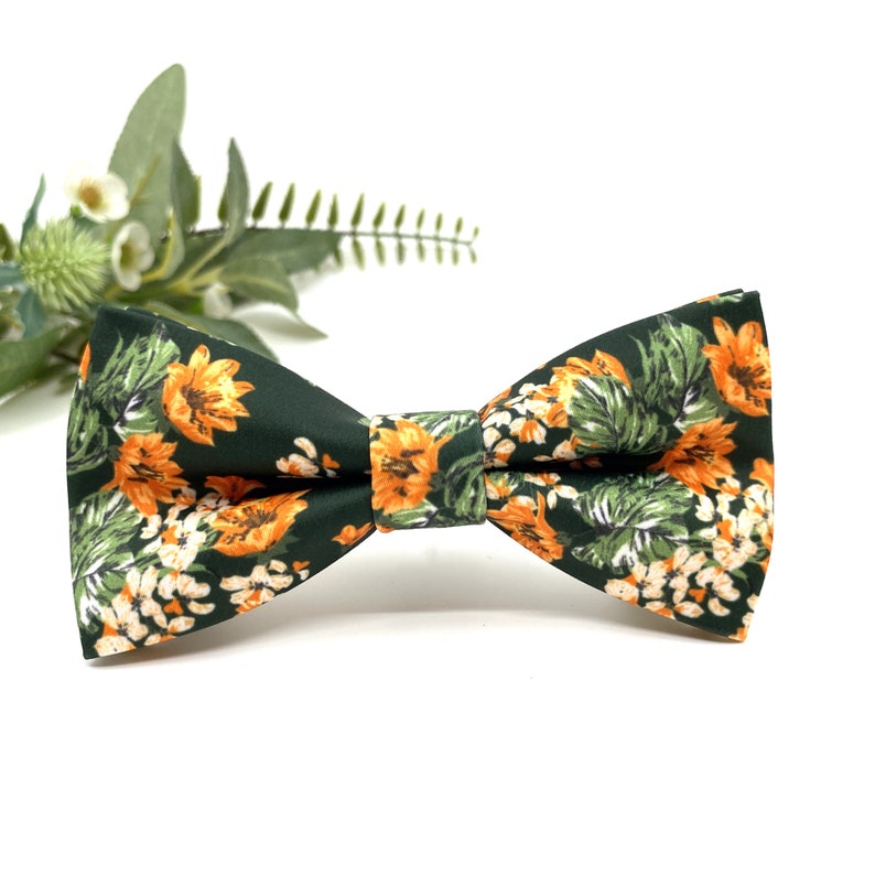 Peut inclure: Un n&oelig;ud papillon vert fonc&eacute; orn&eacute; d'un motif floral vif compos&eacute; de fleurs orange et blanches ainsi que de feuilles vertes. Le tissu pr&eacute;sente une finition lisse et le n&oelig;ud est structur&eacute; par un d&eacute;tail central reliant les deux ailes sym&eacute;triques.