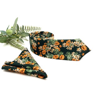 Peut inclure: Une cravate vert fonc&eacute; et une pochette de costume assortie pli&eacute;e, toutes deux orn&eacute;es d'un motif floral vibrant avec des fleurs orange et des feuilles vertes. La cravate est enroul&eacute;e et la pochette est pli&eacute;e en forme de triangle.
