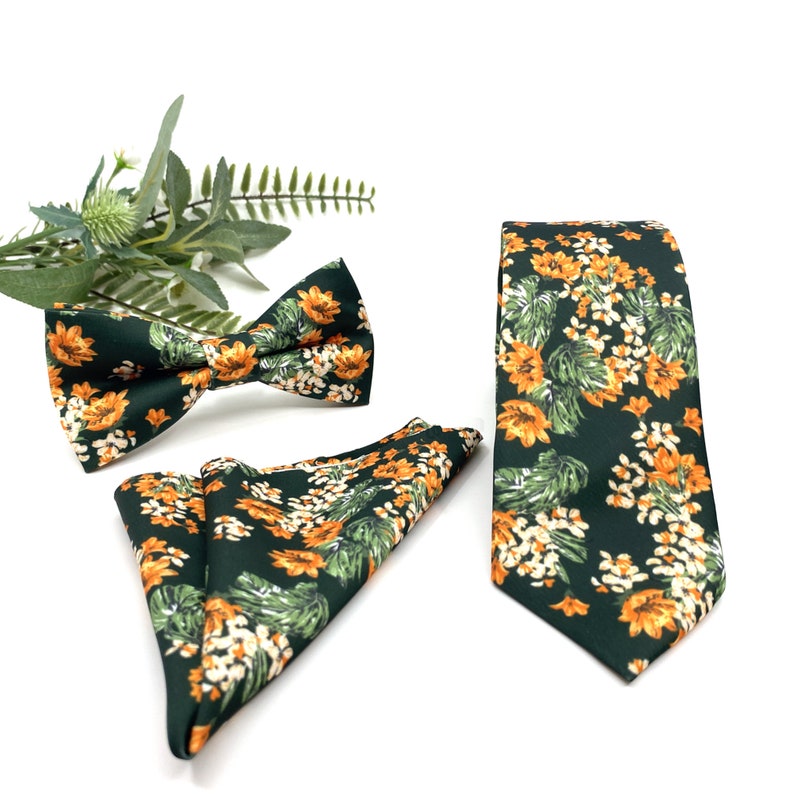Peut inclure: Une cravate, un n&oelig;ud papillon pr&eacute;-nou&eacute; et une pochette pli&eacute;e vert fonc&eacute;, tous orn&eacute;s d'un motif r&eacute;p&eacute;titif de fleurs orange, de petites fleurs blanches et de feuilles vertes. L'ensemble est pr&eacute;sent&eacute; avec un brin de feuillage vert.