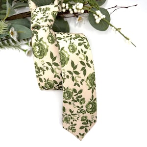 Puede incluir: Una corbata con fondo color crema y un patrón repetitivo de rosas y hojas de color verde oscuro. La corbata está anudada en la parte superior y cuelga hacia abajo, mostrando el estampado floral a lo largo de toda la superficie de la tela.