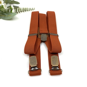 May include: A pair of rust-colored suspenders with brass clips. The suspenders are made of a woven fabric and have a classic design.