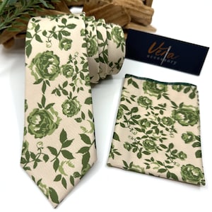 May include: A necktie and a matching pocket square featuring a cream-colored background with a repeating pattern of green roses and leaves. A dark rectangular tag with the text Vera accessory is positioned next to the folded necktie and square.