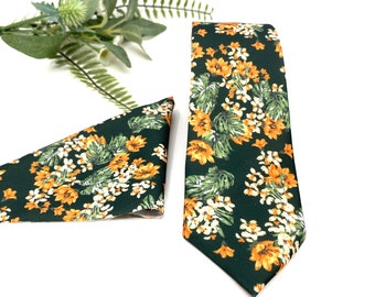 May include: A dark green necktie and a matching triangular pocket square, both featuring a repeating floral pattern of orange lilies, small white blossoms, and green leaves, displayed alongside a sprig of green foliage on a white background.