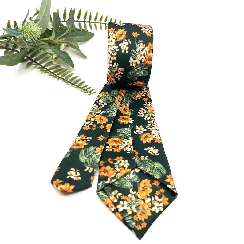 Peut inclure: Une cravate vert fonc&eacute; orn&eacute;e d'un motif r&eacute;p&eacute;titif de fleurs orange vif et de feuilles de monstera tropicales vertes. La cravate est pli&eacute;e sur le dessus, r&eacute;v&eacute;lant la texture du tissu et l'imprim&eacute; floral et botanique d&eacute;taill&eacute; sur toute la surface.