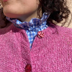Op de afbeelding: Een close-up van een roze gebreid vest met twee rode knopen. Het vest wordt gedragen over een blauw-wit geruit overhemd met kraag. Het vest heeft een levendige roze tint.