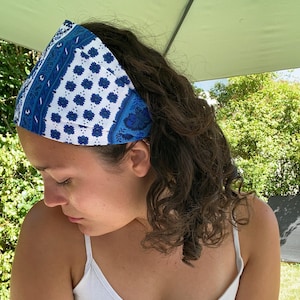 May include: A wide fabric headband featuring a repeating pattern of blue and white floral and geometric motifs. The design includes vertical bands of intricate blue paisley-style details alongside sections of small, dark blue floral shapes.
