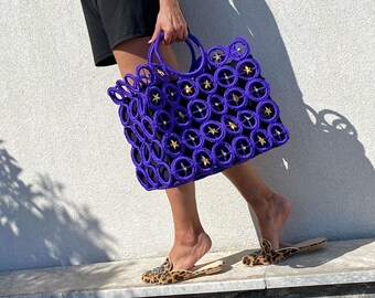 May include: A purple and gold star-shaped tote bag with a woven design. The bag is being held by a person wearing a black dress and leopard print shoes.