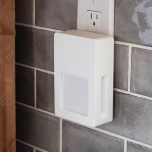 May include: A white, rectangular night light with a square, frosted panel is plugged into an electrical outlet. The night light is mounted on a gray tiled wall. The outlet is white.