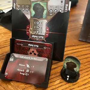 May include: A tabletop board game setup featuring cards and tokens. The cards are in a black holder, with text like "Calculated Strike" and "Black Sludge." A character token stands on a black base. The game pieces are on a wooden surface.