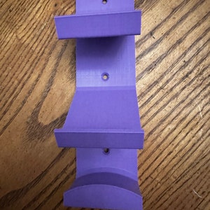 May include: A solid purple, 3D-printed wall mount with three holes for mounting. The mount has two shelves and a curved bottom. The object is made of plastic and is designed to hold an item.