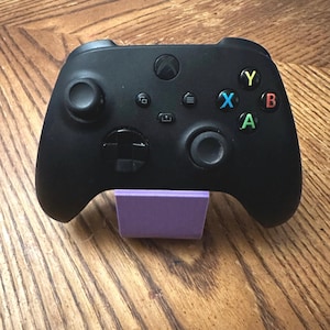 May include: Black Xbox controller with colored buttons (Y, X, B, A) and a purple stand. The controller has two joysticks, a directional pad, and several function buttons. The stand is a solid, rectangular shape.