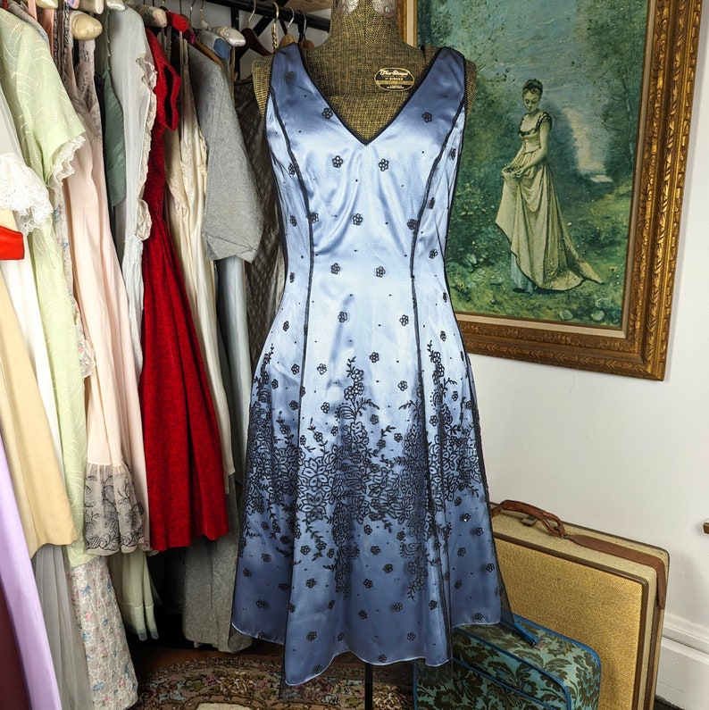 May include: A light blue satin dress with black lace detailing. The dress has a V-neckline and a fitted bodice. The skirt is full and flows out from the waist. The dress is perfect for a special occasion.