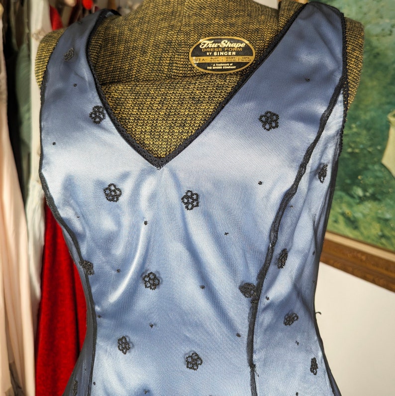 May include: A blue sleeveless dress with black lace trim and a floral pattern. The dress has a V-neckline and is made of a silky fabric. The label on the dress reads "True Shape Dress Form by Singer".