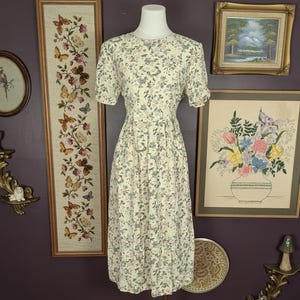 May include: A vintage cream-colored dress with a floral pattern. The dress features short sleeves and a gathered skirt. The dress is displayed on a mannequin. The background includes framed artwork and decorative items.