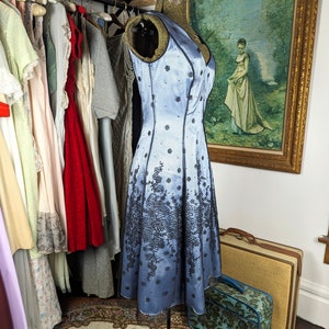 May include: A light blue satin dress with black floral embroidery. The dress has a fitted bodice and a full skirt. It is a vintage style dress.
