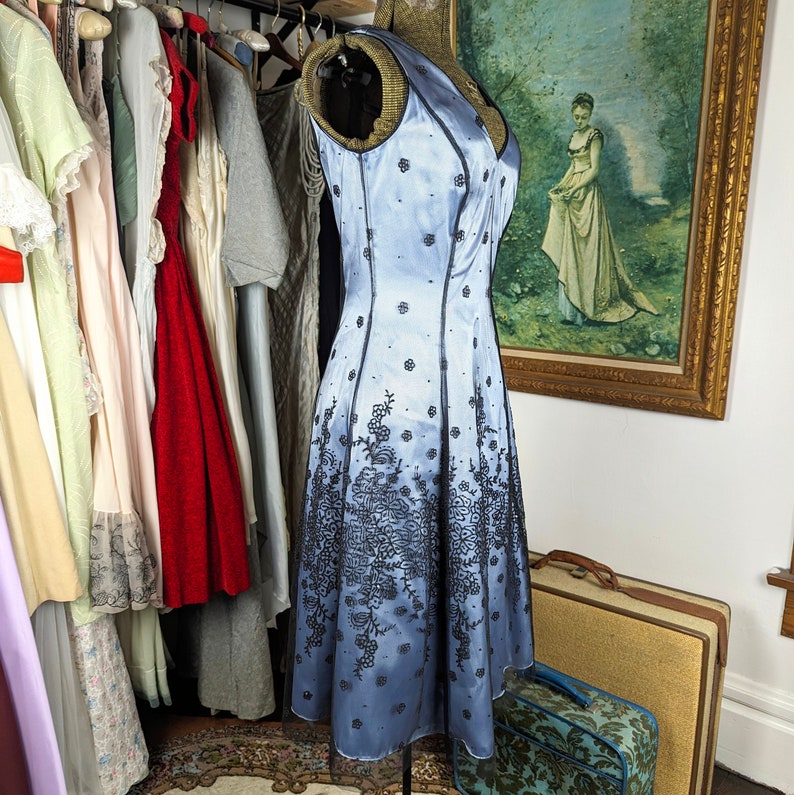 May include: A light blue satin dress with black floral embroidery. The dress has a fitted bodice and a full skirt. It is a vintage style dress.