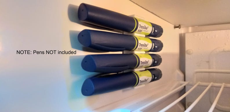 May include: Four blue insulin pens are stored in a white refrigerator door organizer. The pens are labeled "Tresiba" and are stored vertically. The organizer is attached to the refrigerator door with adhesive.