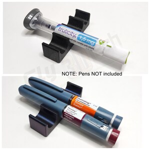 May include: A black 3D printed pen holder with two slots for insulin pens. The holder is shown with two insulin pens, one blue and one orange. The text on the pens reads "Humalog Junior 100 unidades/ml KwikPen" and "Humalog KwikPen".