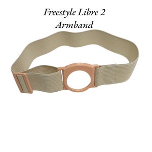 Może przedstawiać: Beżowa elastyczna opaska z okrągłą obudową z tworzywa sztucznego w kolorze brzoskwiniowym na czujnik Freestyle Libre 2. Opaska posiada pasującą klamrę regulacyjną z tworzywa sztucznego. Widoczny jest tekst Freestyle Libre 2 Armband.