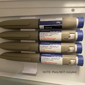 May include: Four brown and blue insulin pens with labels that read "LYUMJEV&trade; KwikPen&reg; (insulin lispro-aabc) Injection For Single Patient Use Only 100 units per ml (U-100)" and "NOC 0002-8207-01". The pens are hanging on a white plastic holder. The text "NOTE: Pens NOT included" is printed below the holder.