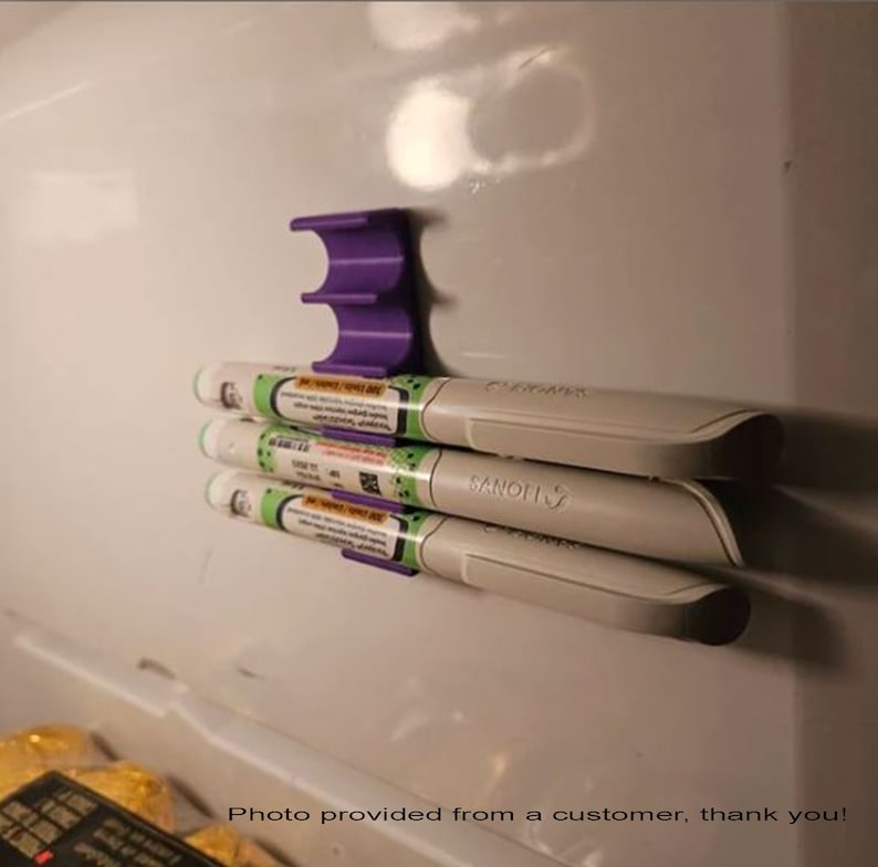 May include: A purple 3D printed wall mount holds three white medical pens. The pens are labeled "Sanofi" and "Clexane".