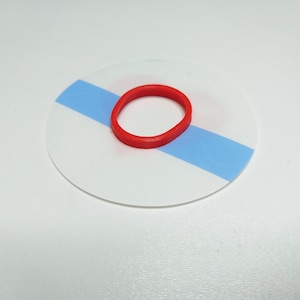 May include: A circular white disc featuring a horizontal light blue stripe across the center, with a small, solid red rubber band resting on top of the disc. The object is flat and circular, displaying a clean, simple design with contrasting colors.