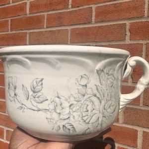 May include: A white ceramic planter with a handle and floral design. The planter features a detailed illustration of roses and leaves in a muted blue-grey colour. The planter is held against a red brick wall.